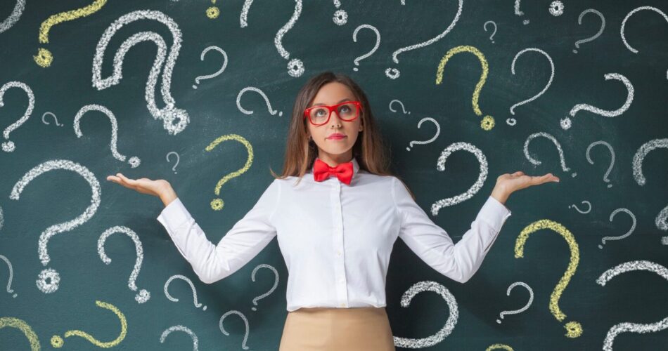 The image features a person standing in front of a chalkboard filled with numerous question marks drawn in white and yellow chalk. The individual is wearing a white shirt with a red bow tie and red glasses, and has their arms raised with palms facing upwards, suggesting a questioning or presenting gesture. The chalkboard is dark green, providing a stark contrast to the bright chalk marks. The person is positioned centrally, with the question marks evenly distributed around them, creating a visually engaging backdrop. The overall composition emphasizes the theme of inquiry and uncertainty.