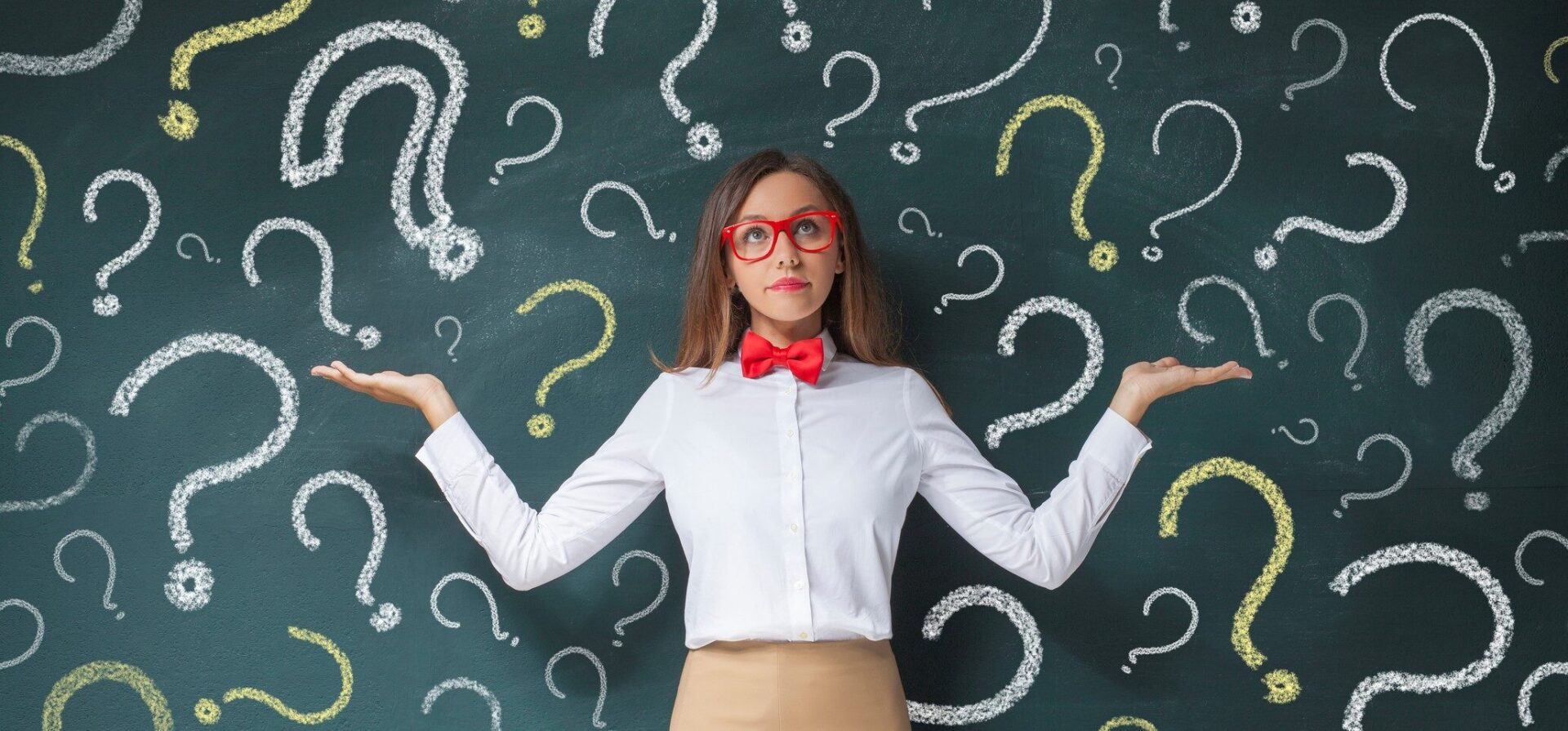 The image features a person standing in front of a chalkboard filled with numerous question marks drawn in white and yellow chalk. The individual is wearing a white shirt with a red bow tie and red glasses, and has their arms raised with palms facing upwards, suggesting a questioning or presenting gesture. The chalkboard is dark green, providing a stark contrast to the bright chalk marks. The person is positioned centrally, with the question marks evenly distributed around them, creating a visually engaging backdrop. The overall composition emphasizes the theme of inquiry and uncertainty.
