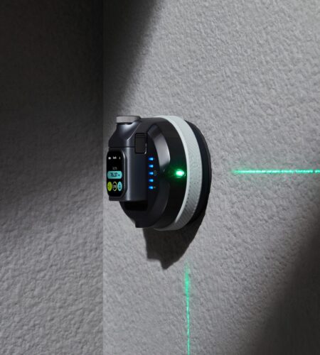 The image shows a wall-mounted laser level device. The device is black with a circular shape and a digital display on the front. The display shows a measurement reading of "76.53" and has several buttons below it, including a green button and a yellow button. There are also three blue indicator lights on the side of the device. A green laser beam is visible, projecting a horizontal line on the wall, indicating the device's function. The wall is textured and gray, and the device is mounted at a corner, suggesting it is used for leveling or alignment purposes.