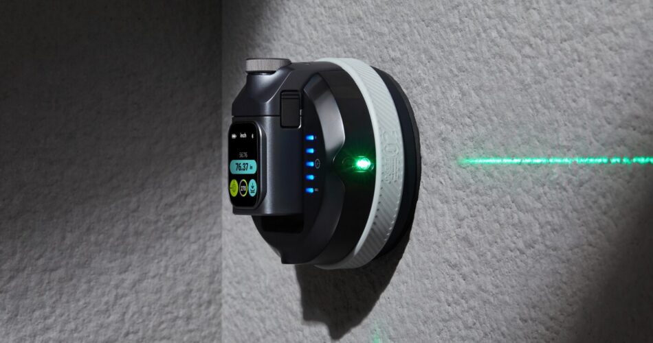 The image shows a wall-mounted laser level device. The device is black with a circular shape and a digital display on the front. The display shows a measurement reading of "76.53" and has several buttons below it, including a green button and a yellow button. There are also three blue indicator lights on the side of the device. A green laser beam is visible, projecting a horizontal line on the wall, indicating the device's function. The wall is textured and gray, and the device is mounted at a corner, suggesting it is used for leveling or alignment purposes.