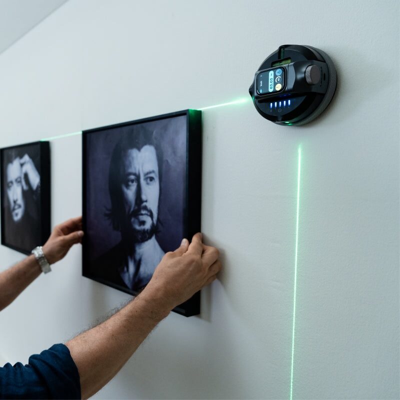 The image shows a person adjusting a framed picture on a white wall. The individual is wearing a dark blue long-sleeve shirt and a silver wristwatch. A circular device mounted on the wall emits a green laser line, which is being used to align the picture. The device has a digital display and several blue indicator lights. The picture being adjusted is a black and white portrait, and there is another similar picture to the left. The laser line is used to ensure the picture is straight and properly aligned with the others.