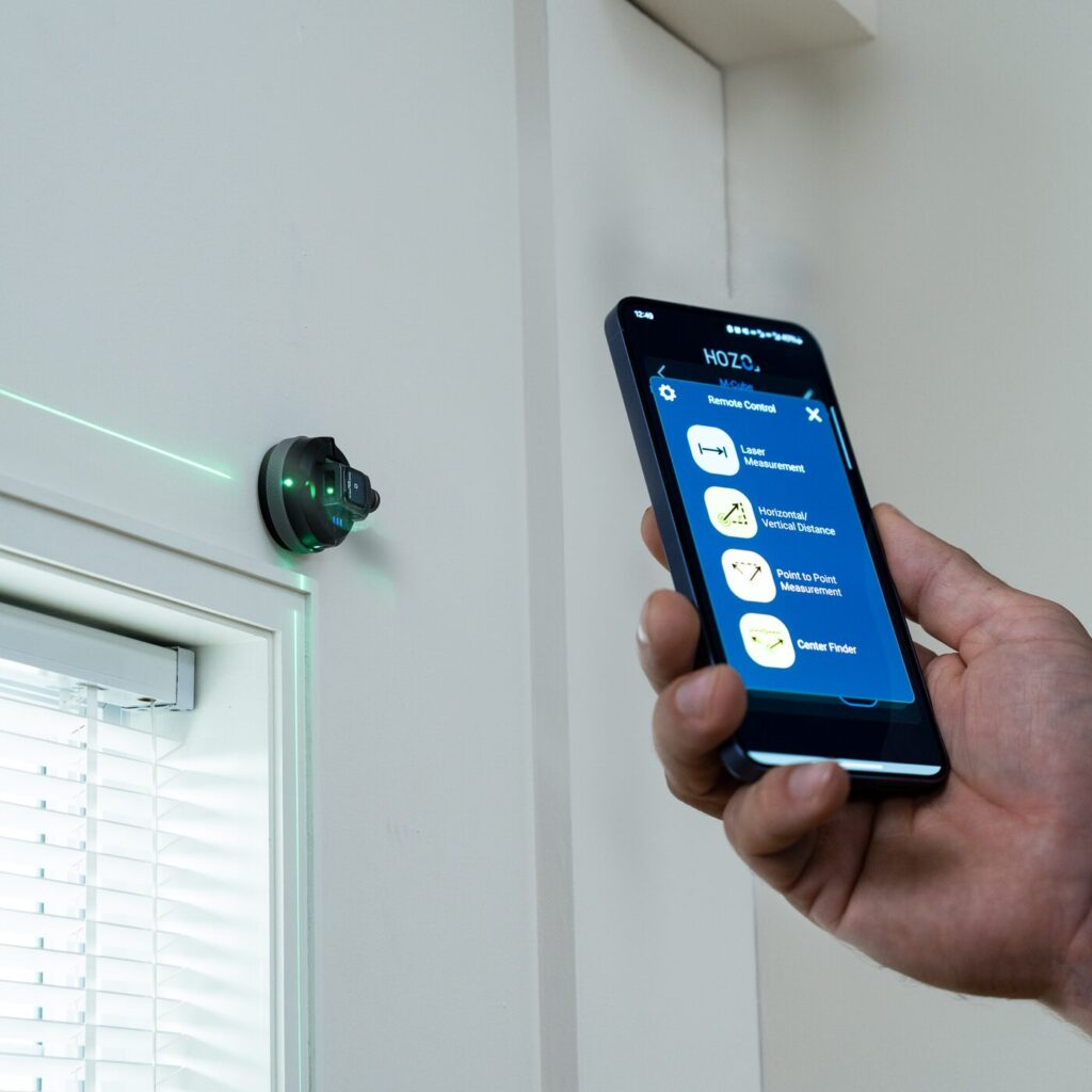The image shows a close-up of a hand holding a smartphone displaying an app interface. The app is titled "HOZO" and offers several features: "Remote Control," "Laser Measurement," "Horizontal/Vertical Distance," "Point to Point Measurement," and "Center Finder." The smartphone is held in front of a wall-mounted device emitting a green laser line, which is used for measuring distances. The device is circular with a black body and green lights, mounted on a white wall near a window with partially open blinds. The laser line is visible on the wall, indicating the device's function. The smartphone screen is blue with white text and icons, and the time displayed is 12:49.
