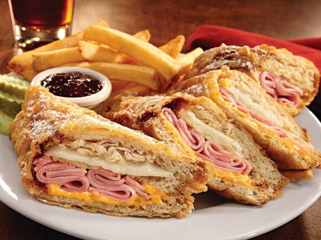 The image features a plate of food with a sandwich and side items. The sandwich is cut into four triangular pieces, revealing layers of ham, melted cheese, and possibly shredded chicken, all encased in a golden-brown, flaky crust. The sandwich is dusted with a light coating of powdered sugar, giving it a slightly sweet appearance. Accompanying the sandwich are a portion of golden French fries and a small white ramekin containing a dark, chunky sauce, likely a barbecue or sweet chili sauce. A pickle spear is also present on the plate, adding a touch of green. In the background, there is a glass of a dark beverage, possibly soda, and a red napkin, suggesting a casual dining setting. The plate is set on a wooden table, enhancing the warm and inviting atmosphere of the meal.