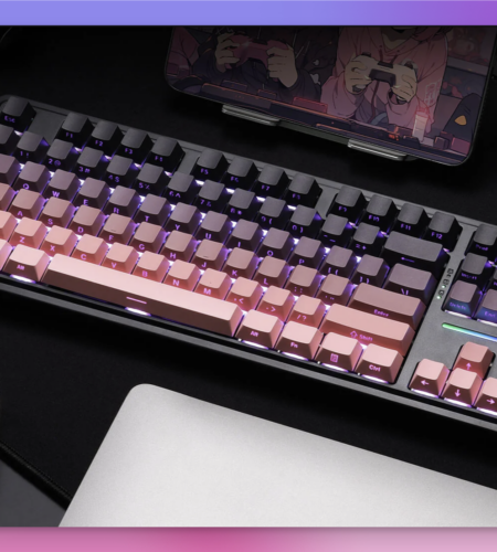 The image features a mechanical keyboard with a gradient keycap design, transitioning from black to pink, with purple backlighting. The keyboard is placed on a black desk mat, with a transparent game controller to the left and a silver laptop to the right. A tablet displaying an anime scene is propped up in the background. A small astronaut figurine is visible on the right side of the keyboard. The overall setup suggests a gaming or creative workspace.