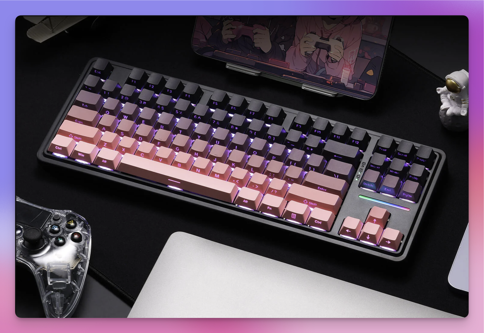 The image features a mechanical keyboard with a gradient keycap design, transitioning from black to pink, with purple backlighting. The keyboard is placed on a black desk mat, with a transparent game controller to the left and a silver laptop to the right. A tablet displaying an anime scene is propped up in the background. A small astronaut figurine is visible on the right side of the keyboard. The overall setup suggests a gaming or creative workspace.