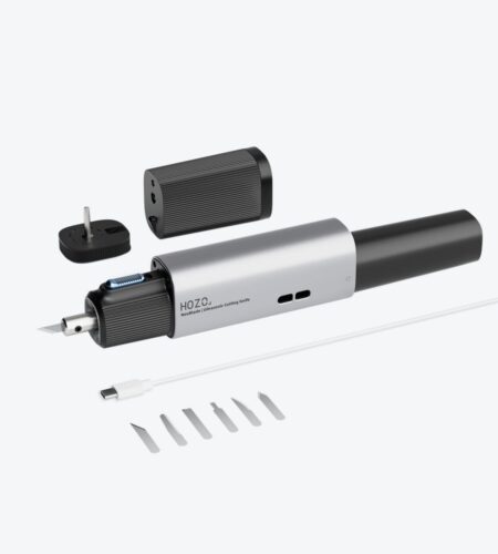 A product shot displays a silver-colored, pen-shaped device with a plug adapter and a set of replacement blades lying nearby on a white surface. The device features a textured grip section in the middle and a pointed nib at one end. To the right of the device, a small plastic tray holds a series of thin, silver-colored replacement blades, appearing identical in shape and size. A black plug adapter, presumably for powering the device, is positioned at the upper left corner of the frame. The text “HOZO” and “ReciProCut | Ultrasonic Cutting Knife” is visible on the device’s body.