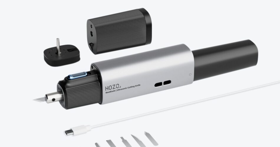 A product shot displays a silver-colored, pen-shaped device with a plug adapter and a set of replacement blades lying nearby on a white surface. The device features a textured grip section in the middle and a pointed nib at one end. To the right of the device, a small plastic tray holds a series of thin, silver-colored replacement blades, appearing identical in shape and size. A black plug adapter, presumably for powering the device, is positioned at the upper left corner of the frame. The text “HOZO” and “ReciProCut | Ultrasonic Cutting Knife” is visible on the device’s body.
