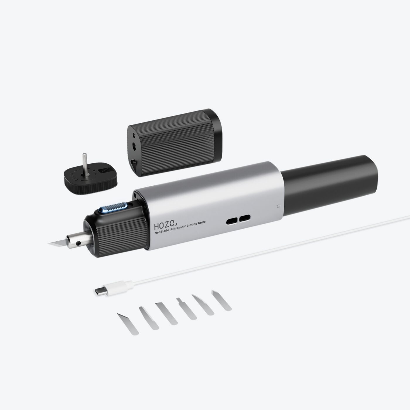 A product shot displays a silver-colored, pen-shaped device with a plug adapter and a set of replacement blades lying nearby on a white surface. The device features a textured grip section in the middle and a pointed nib at one end. To the right of the device, a small plastic tray holds a series of thin, silver-colored replacement blades, appearing identical in shape and size. A black plug adapter, presumably for powering the device, is positioned at the upper left corner of the frame. The text “HOZO” and “ReciProCut | Ultrasonic Cutting Knife” is visible on the device’s body.