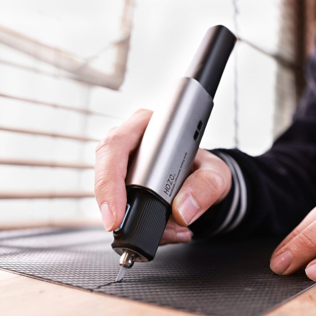 A hand is holding a gray pen-shaped tool over a dark, patterned surface. The tool has a ribbed section near the tip and a smooth, cylindrical body that widens toward the back. The tip appears to be metallic and pointed, likely for detailed work. The hand is partially visible, with fingers wrapped around the tool’s body. The surface below is a dark gray with a grid-like pattern of small squares. 