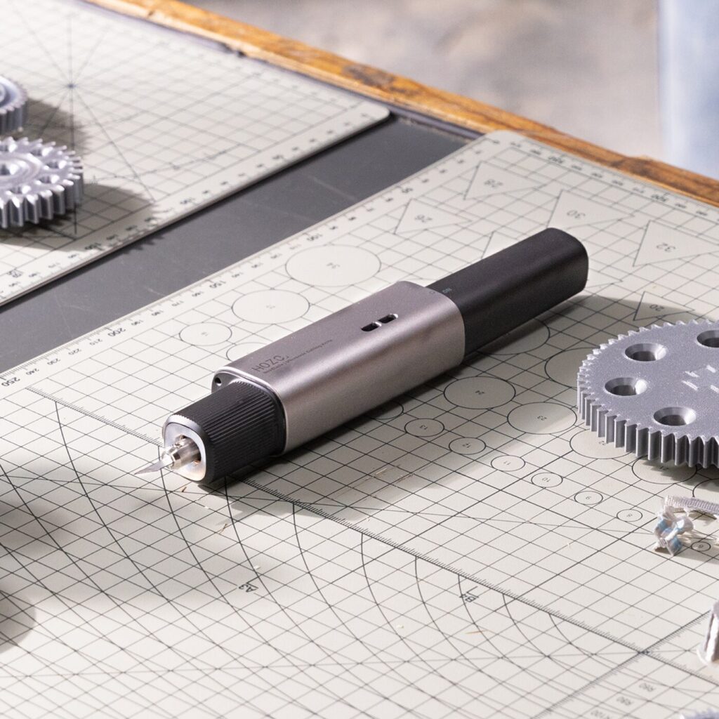 A cylindrical, dark gray device rests diagonally on a light gray surface with a grid pattern. The device has a textured grip section in the middle and smoother sections at both ends. One end features a silver-colored connector, while the other is a solid, rounded cap. Several metallic gears of varying sizes are partially visible in the blurred background, positioned to the left and right of the device. The surface beneath the device is marked with a precise grid, and faint lines radiate outwards from a central point. The text “4020” is visible on the body of the device.