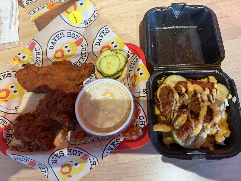 A top-down shot displays a fast-food meal arranged on a light-colored wood-grain surface. The meal consists of a container of fried chicken tenders, a small cup of dipping sauce, two pickle slices, and a black container filled with french fries. The chicken tenders are arranged on small, square paper wrappers with the text “DAVES HOT CHICKEN” and “MEDIUM” printed on them. The dipping sauce is a light orange color in a clear plastic cup. The black container of french fries is full, with visible seasoning on top.