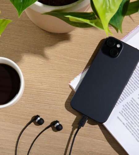 A high-angle, flat-lay shot captures a wooden desk surface arranged with several objects. In the upper section, vibrant green leaves from a potted plant extend from a speckled cream-colored ceramic pot. To the left, a white mug filled with dark coffee sits on the wood grain. On the right side, a black smartphone rests diagonally across an open book with printed text. At the bottom, a pair of black wired earbuds lies on the table, with one wire plugged directly into the bottom port of the phone.