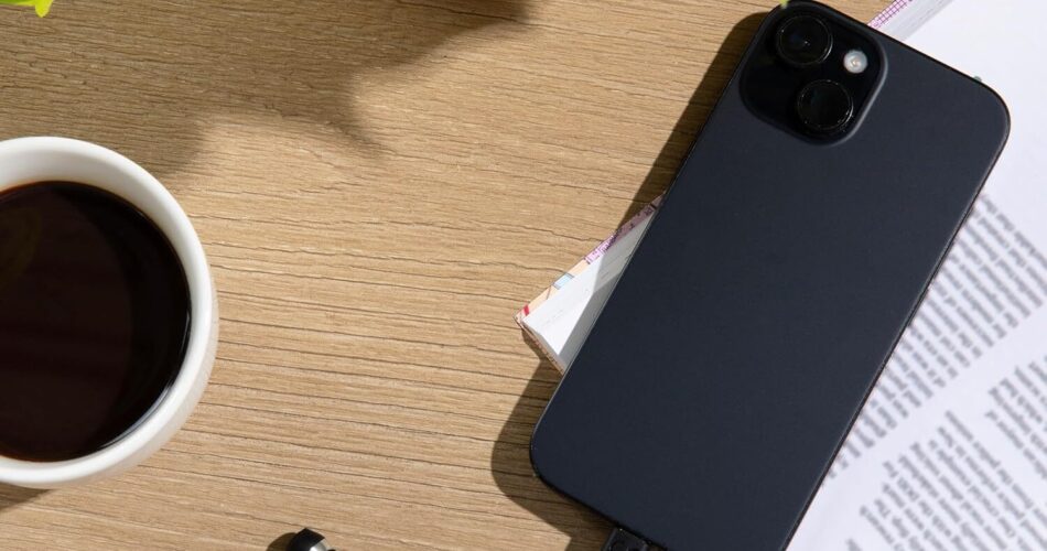 A high-angle, flat-lay shot captures a wooden desk surface arranged with several objects. In the upper section, vibrant green leaves from a potted plant extend from a speckled cream-colored ceramic pot. To the left, a white mug filled with dark coffee sits on the wood grain. On the right side, a black smartphone rests diagonally across an open book with printed text. At the bottom, a pair of black wired earbuds lies on the table, with one wire plugged directly into the bottom port of the phone.