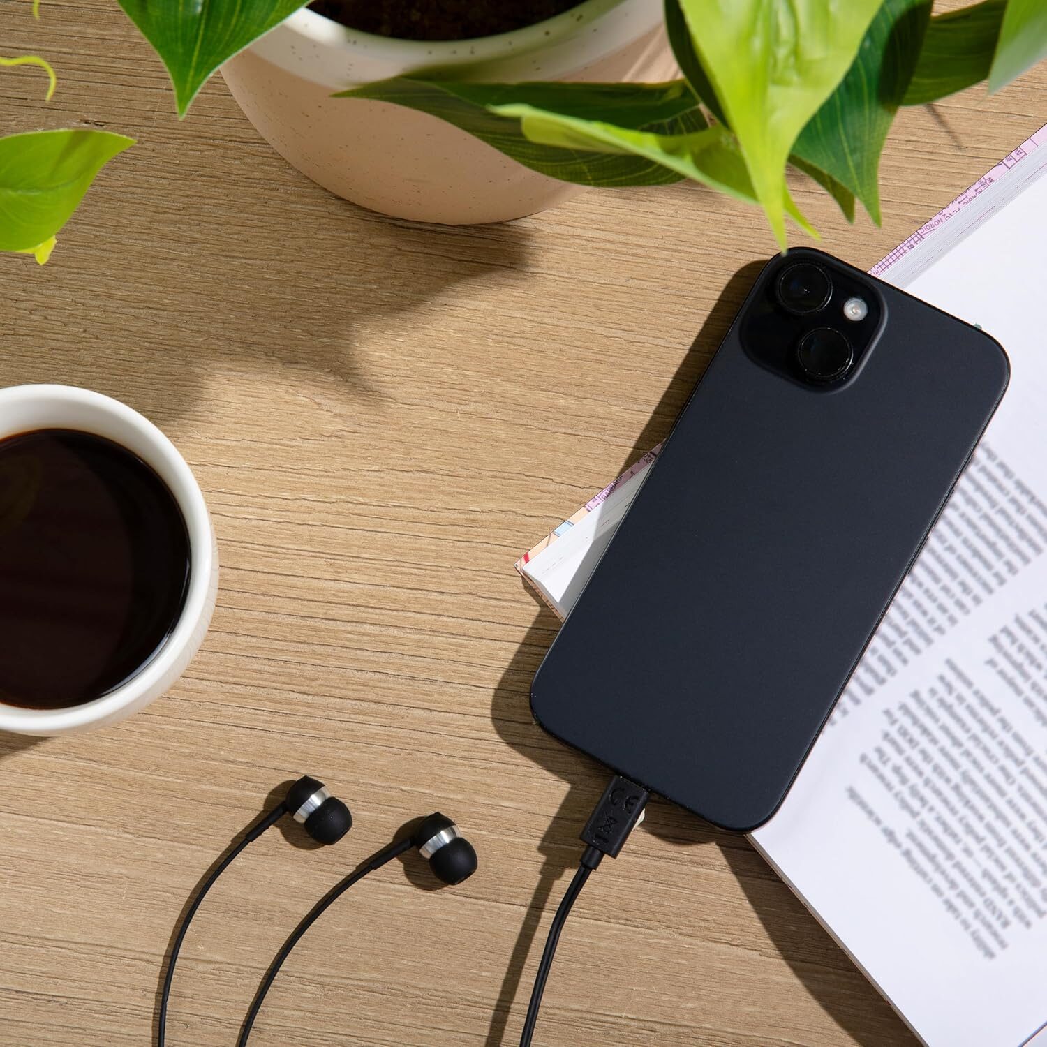A high-angle, flat-lay shot captures a wooden desk surface arranged with several objects. In the upper section, vibrant green leaves from a potted plant extend from a speckled cream-colored ceramic pot. To the left, a white mug filled with dark coffee sits on the wood grain. On the right side, a black smartphone rests diagonally across an open book with printed text. At the bottom, a pair of black wired earbuds lies on the table, with one wire plugged directly into the bottom port of the phone.